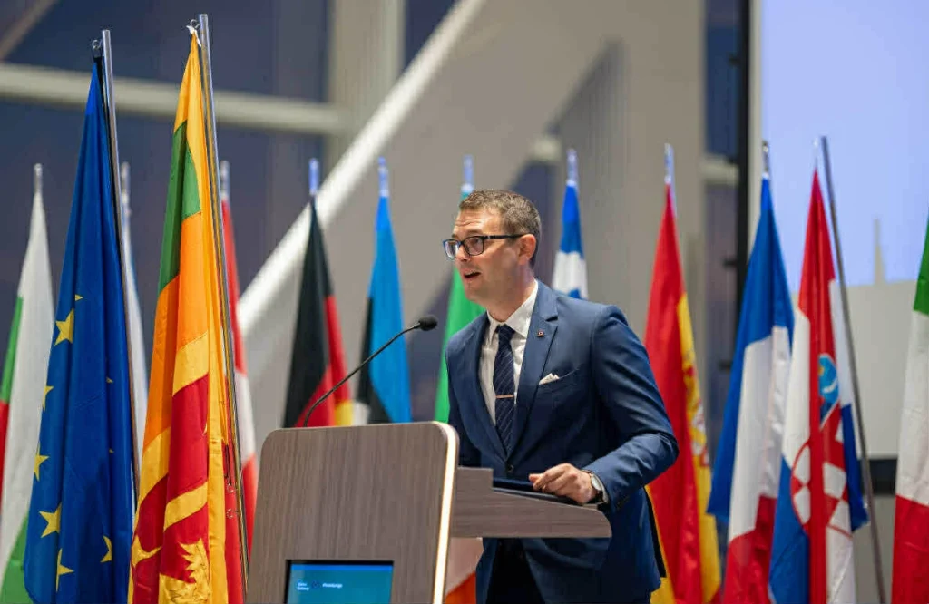 Előadó beszél pulpitusnál, háttérben nemzetközi zászlók