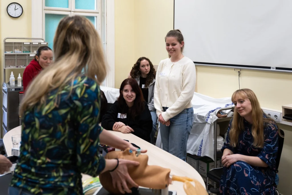 Elsősegély oktatás: instruktor babamanökennel mutat be gyakorlatot, diákok figyelnek